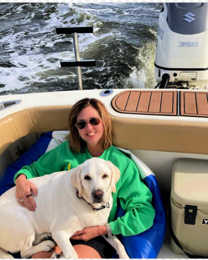 A woman in a green sweatshirt sitting on a boat with a yellow Labrador retriever.