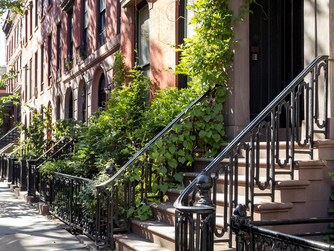 Street view of brownstone buildings with greenery and iron railings.